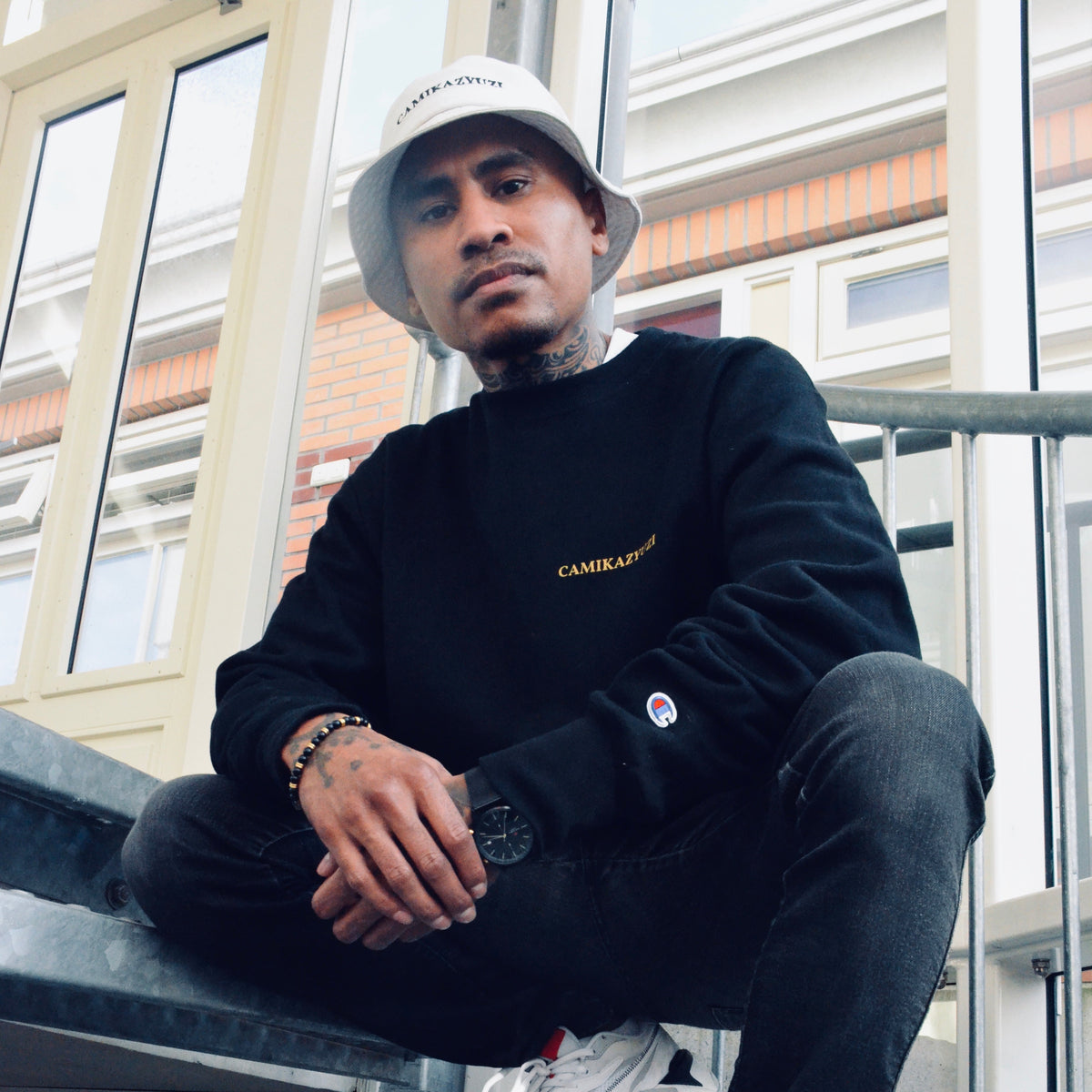 A man wearing a black crewneck sweatshirt with gold 'CAMIKAZYUZI' text on the chest, black jeans, white sneakers, and a white bucket hat, sitting on outdoor metal stairs.