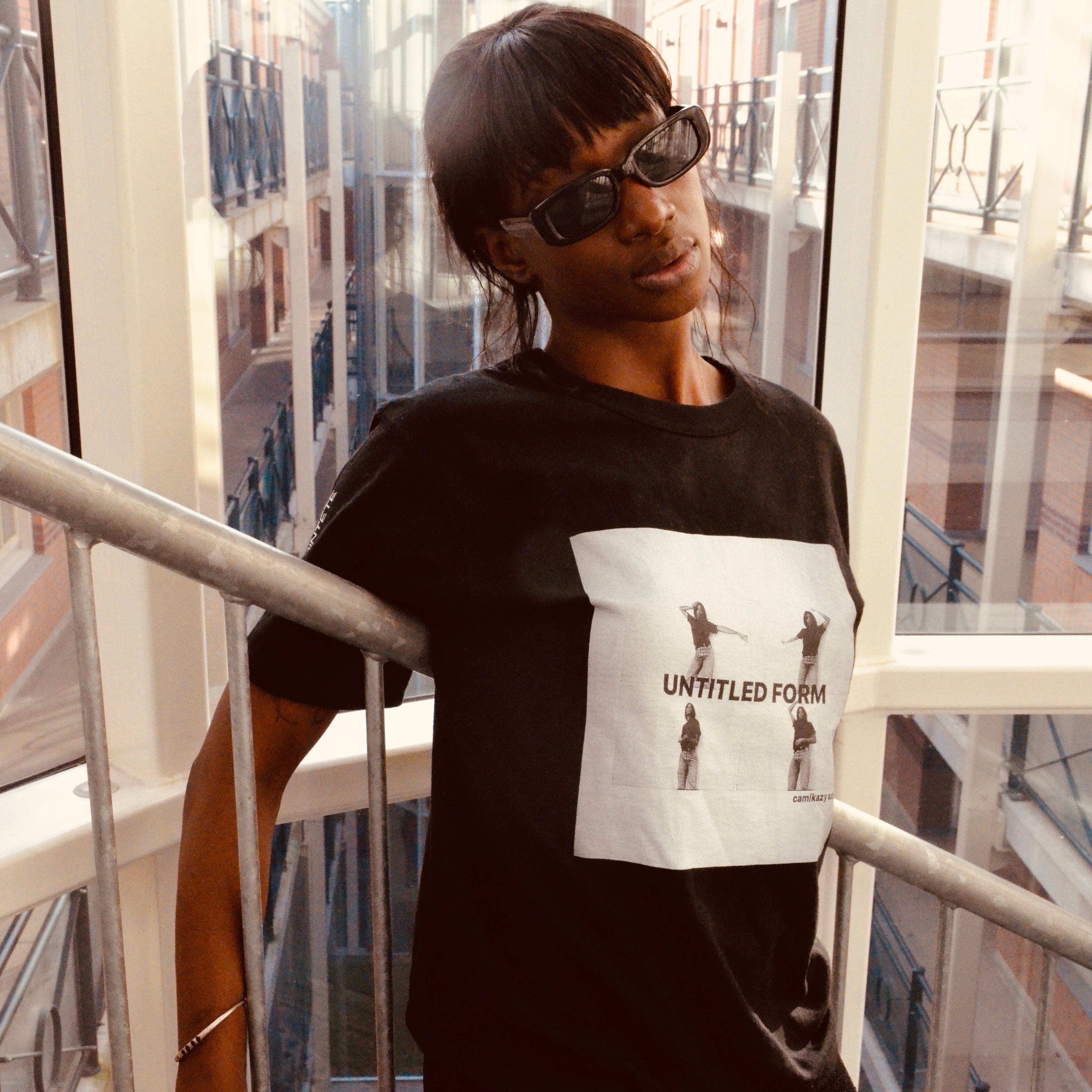 A person wearing a black graphic t-shirt and black pants, leaning on a metal railing in front of large windows with an urban courtyard view. The t-shirt features a white rectangular graphic with the text 'UNTITLED FORM' and small figures.