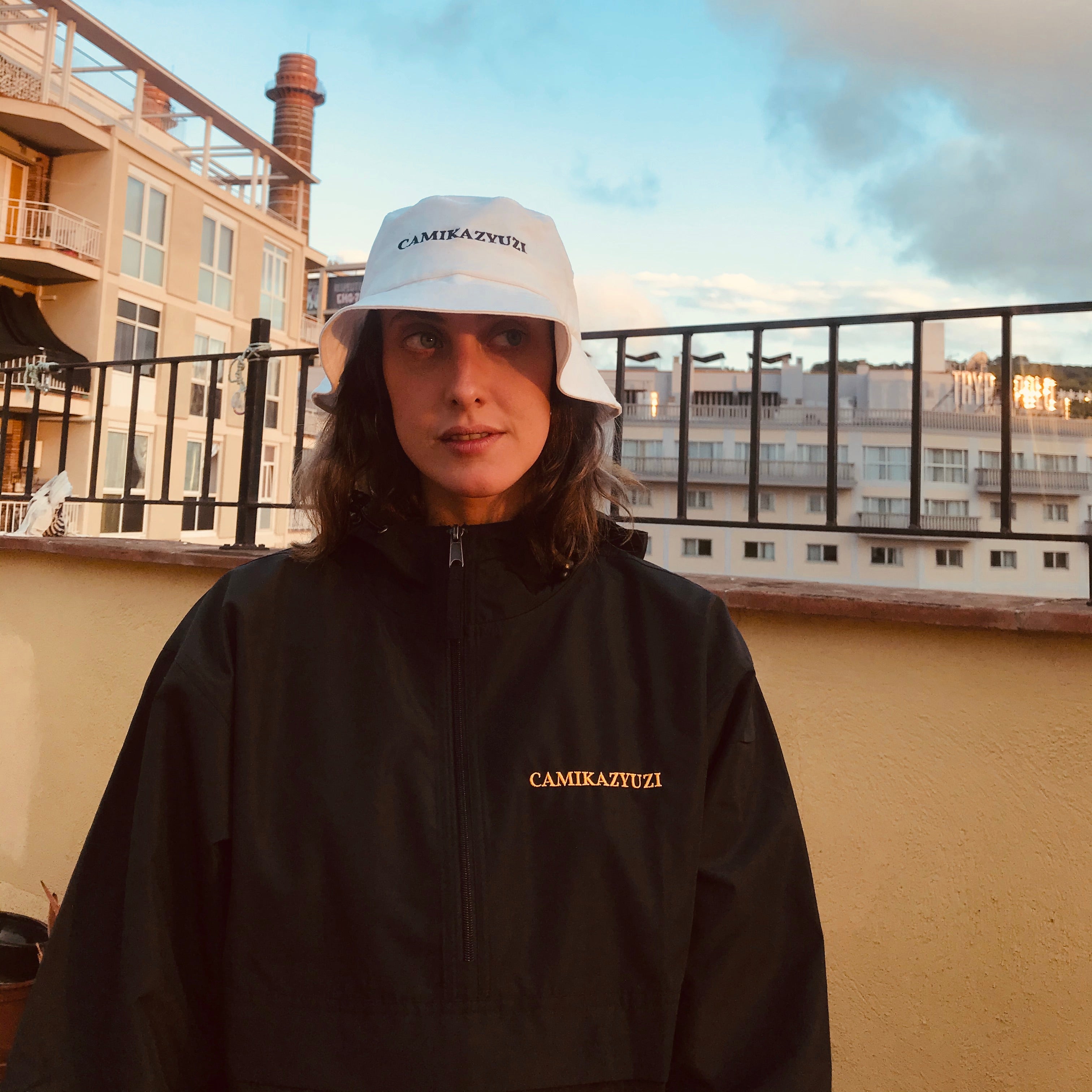 A person wearing a white bucket hat with 'CAMIKAZY UZI' printed on the front and a black zip-up jacket with the same brand name in gold on the chest, standing outdoors on a balcony with buildings and a cloudy sky in the background.